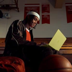 A person in sportswear sits on gym bleachers with a yellow laptop, a basketball, and an orange backpack nearby. Red championship banners hang on the wall in the background.