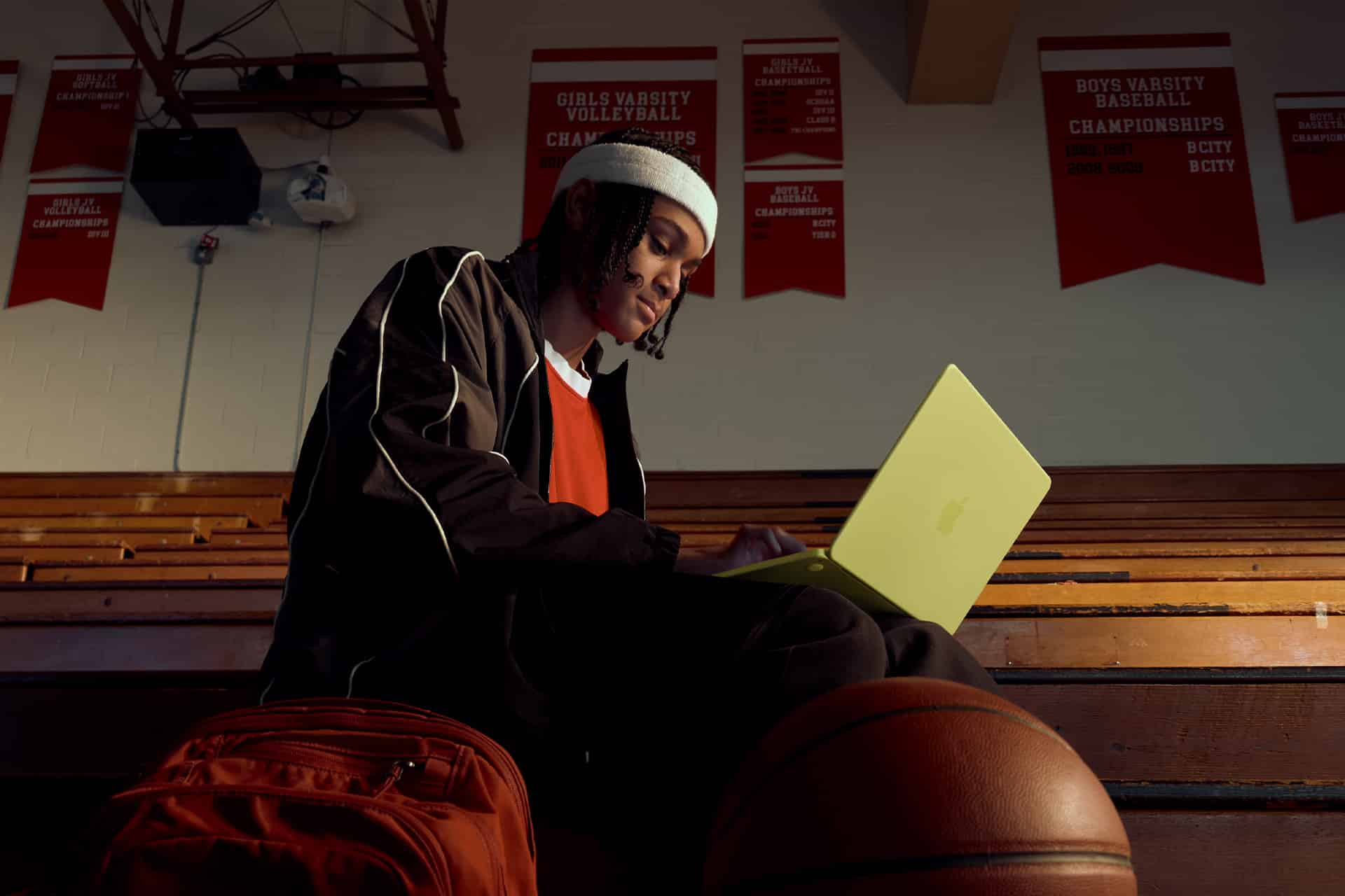 A person in sportswear sits on gym bleachers with a yellow laptop, a basketball, and an orange backpack nearby. Red championship banners hang on the wall in the background.