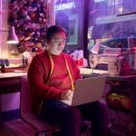 A person in a red sweater with a yellow measuring tape around their neck sits in a sewing studio, working on a laptop. Spools of thread, a sewing machine, and a window with rainy streets are visible in the background.