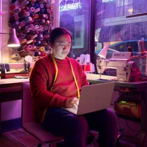 A person in a red sweater with a yellow measuring tape around their neck sits in a sewing studio, working on a laptop. Spools of thread, a sewing machine, and a window with rainy streets are visible in the background.