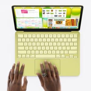A person with rings on their fingers types on a yellow MacBook Neo, its screen displaying multiple open windows against a white background.