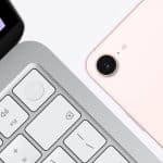 Close-up of a silver laptop keyboard beside the back of a light pink smartphone with camera lens and flash, both on a white surface—an everyday scene reflecting the subtle impact of Apple ecosystem lock in.