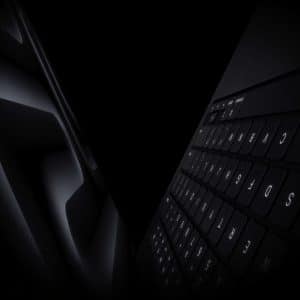 A dark, dramatic close-up of a sleek Apple laptop keyboard and trackpad, with the Apple logo faintly visible in the corner.