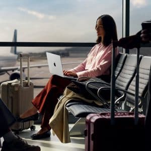 A woman sits in an airport terminal with a MacBook Air M5 on her lap, surrounded by suitcases. She faces a large window overlooking the tarmac, where sunlight streams in and a plane waits outside.