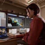 A woman sits at a desk in an office, working on 3D modeling with colorful sculpted figures displayed on a laptop and two monitors. She is focused on her work, surrounded by large windows and modern decor.