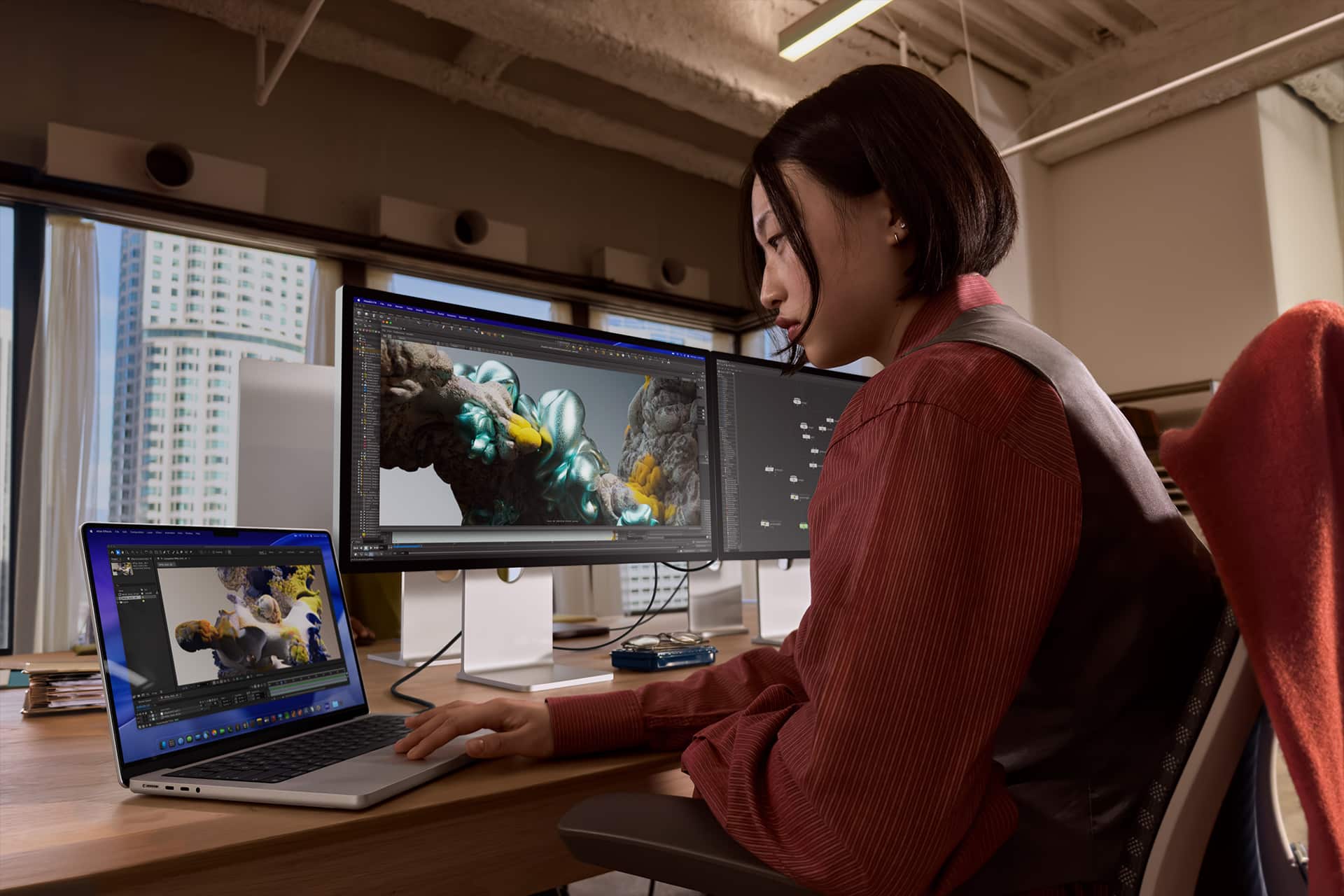 Neo vs Air vs Pro - A woman sits at a desk in an office, working on 3D modeling with colorful sculpted figures displayed on a laptop and two monitors. She is focused on her work, surrounded by large windows and modern decor.
