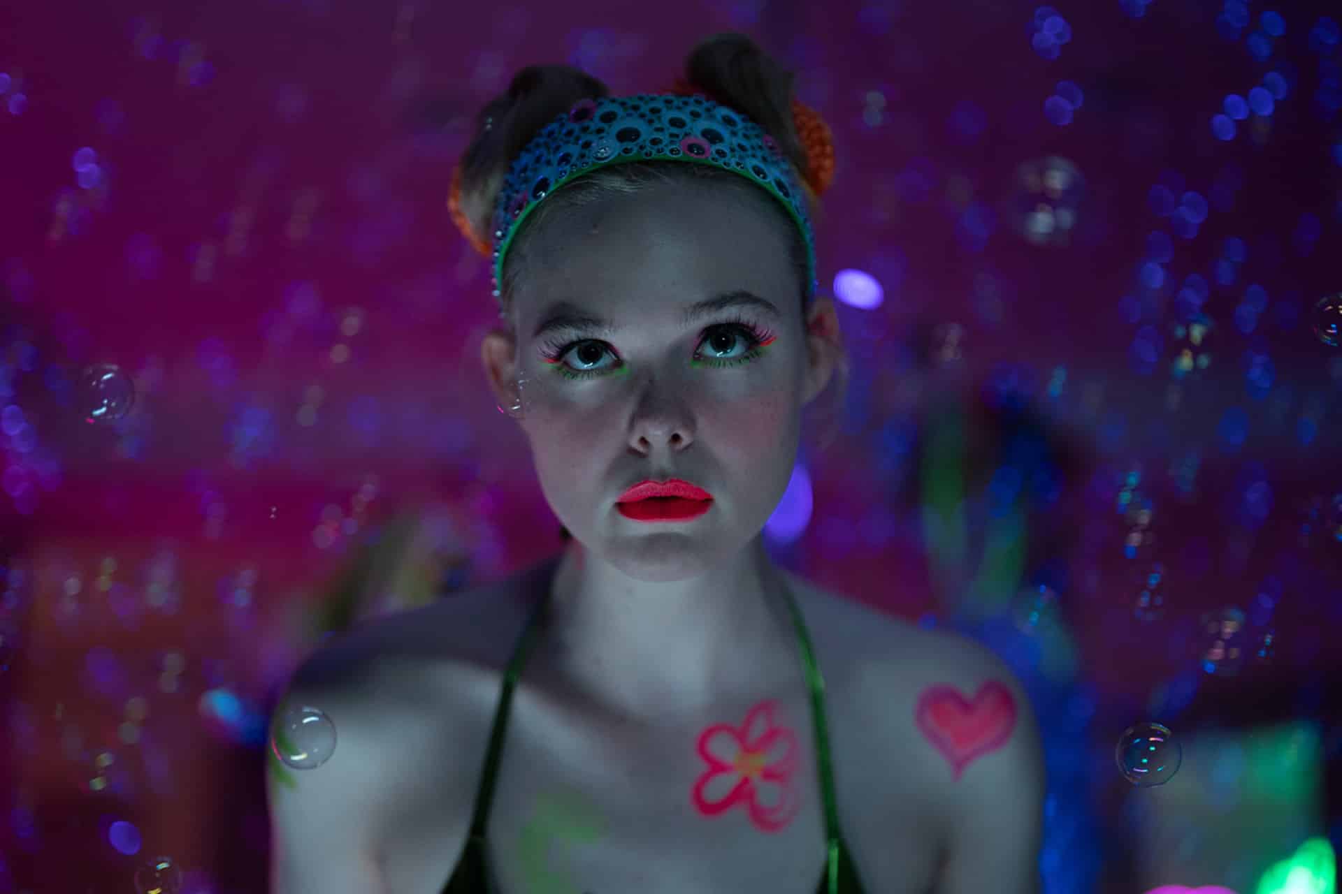 A young person with bright makeup, face gems, and neon body paint looks upward; they wear a jeweled headband and are surrounded by bubbles and colorful, dreamy lighting.