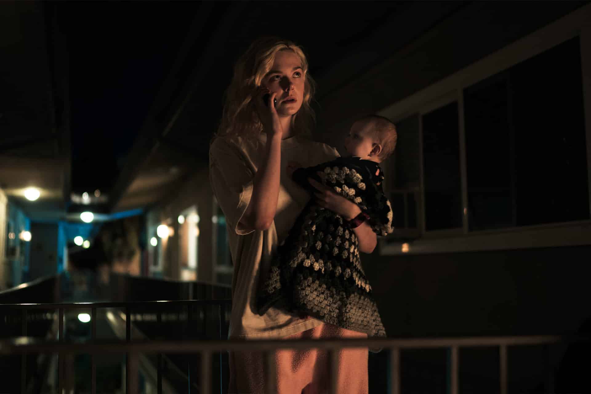 Margo’s Got Money Troubles -A woman stands outside at night holding a baby wrapped in a blanket. She looks concerned while talking on a phone, illuminated by dim outdoor lighting, with apartment buildings in the background.