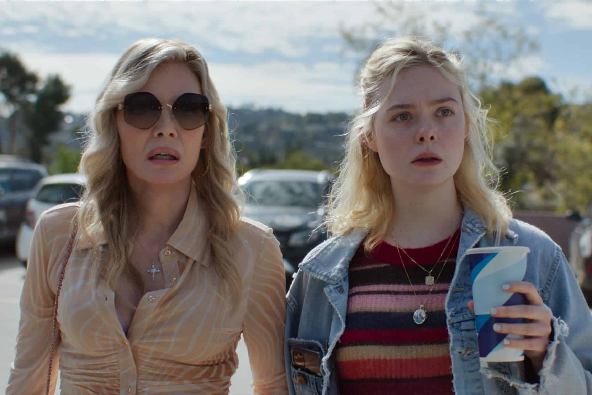 Two women stand outdoors in a parking lot. One wears sunglasses, a striped blouse, and jewelry; the other has blond hair, a denim jacket, a striped sweater, and holds a blue-and-white cup. Both have serious expressions.