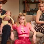 Margo’s Got Money Troubles - Three adults and a baby sit on a couch. A bearded man in black holds the baby, a young woman in a pink top stares ahead, and a woman in a leopard print dress looks at her phone. The background shows shelves and toys.