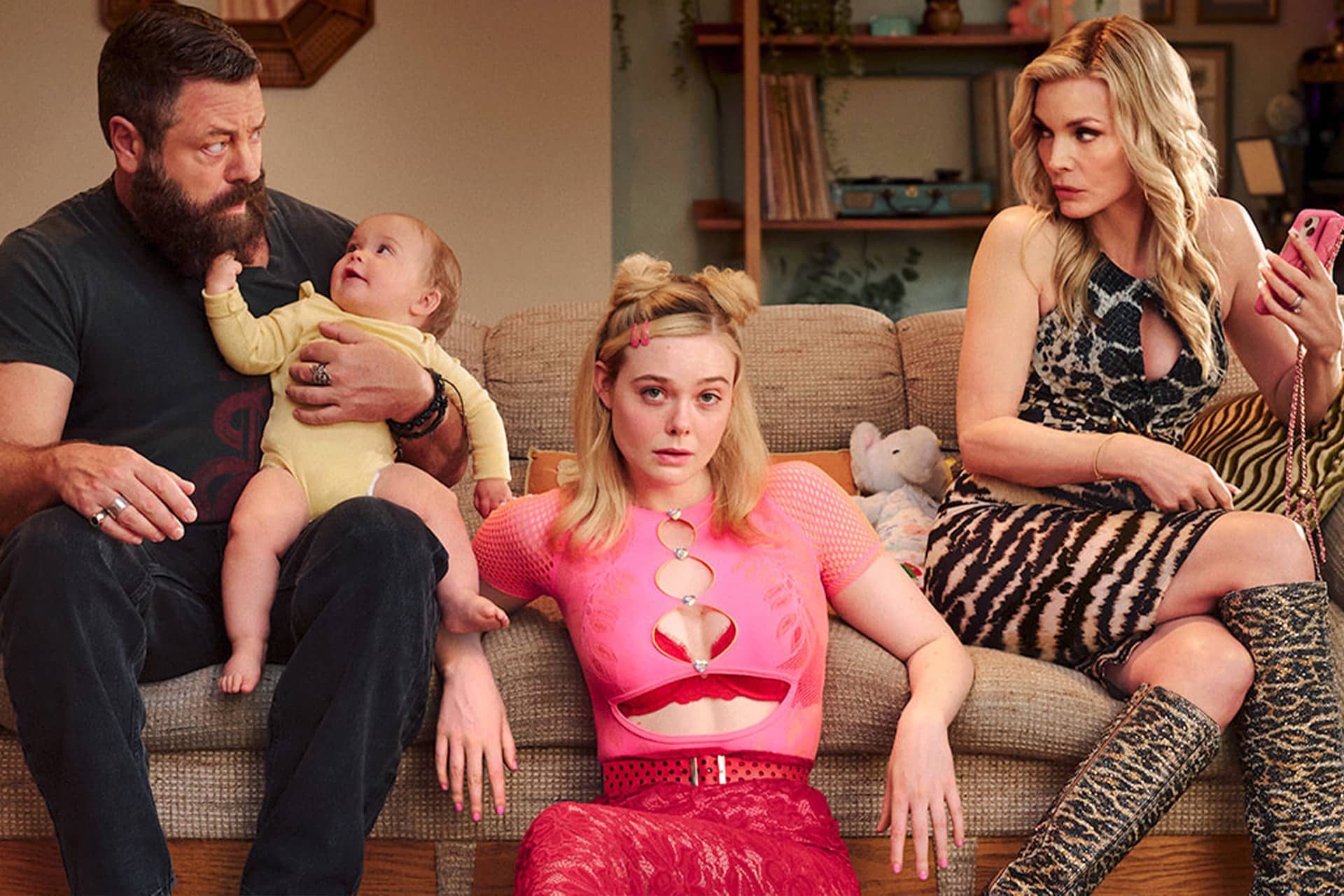Margo’s Got Money Troubles - Three adults and a baby sit on a couch. A bearded man in black holds the baby, a young woman in a pink top stares ahead, and a woman in a leopard print dress looks at her phone. The background shows shelves and toys.