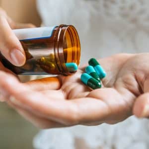 A person pours blue and green capsules from a brown pill bottle into their hand, with a white patterned shirt visible in the background—an everyday scene that highlights the importance of medication reminders.