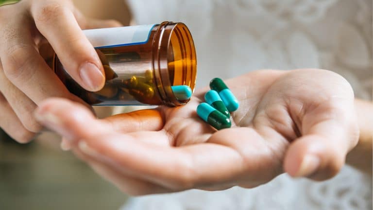 A person pours blue and green capsules from a brown pill bottle into their hand, with a white patterned shirt visible in the background—an everyday scene that highlights the importance of medication reminders.