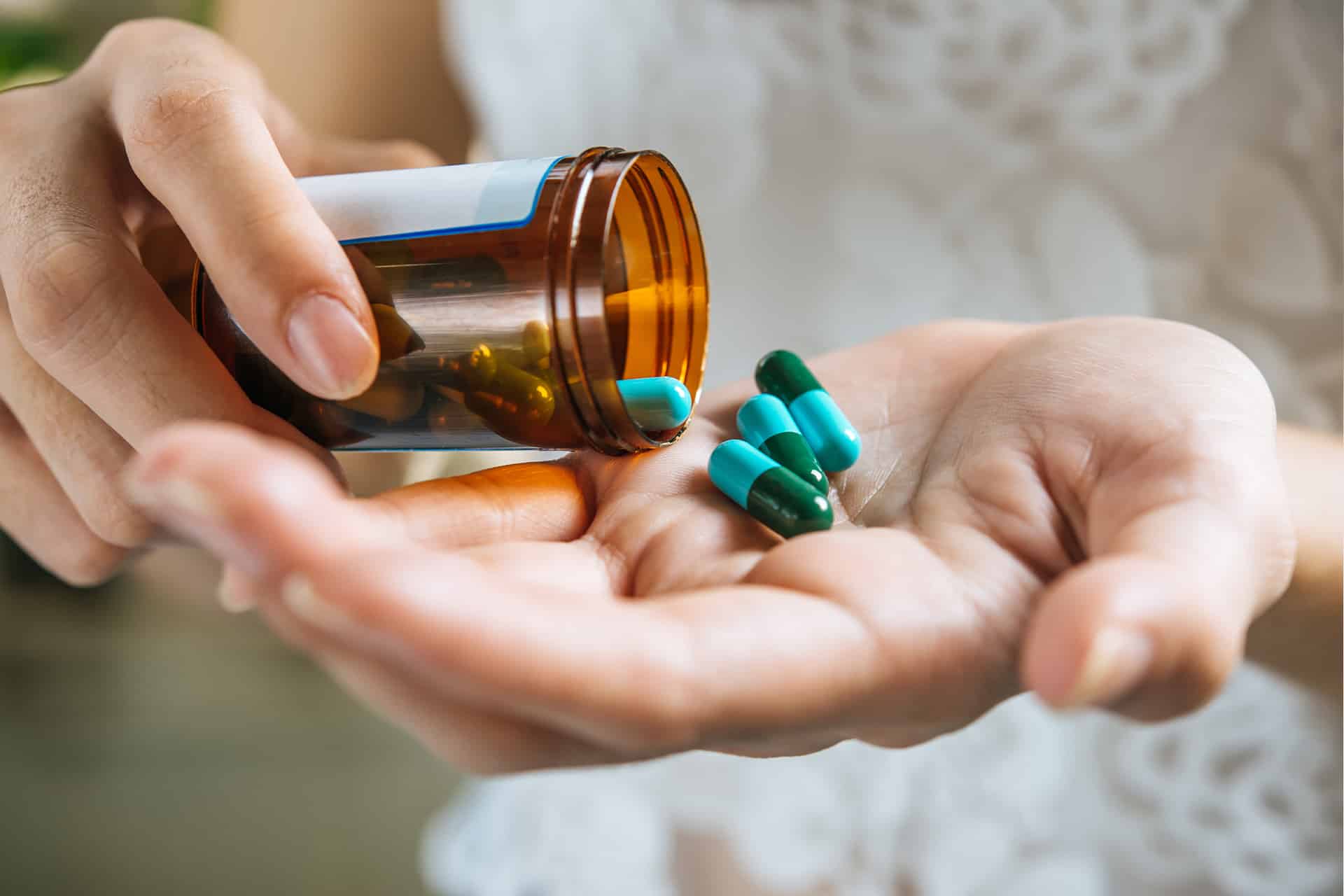 A person pours blue and green capsules from a brown pill bottle into their hand, with a white patterned shirt visible in the background—an everyday scene that highlights the importance of medication reminders.