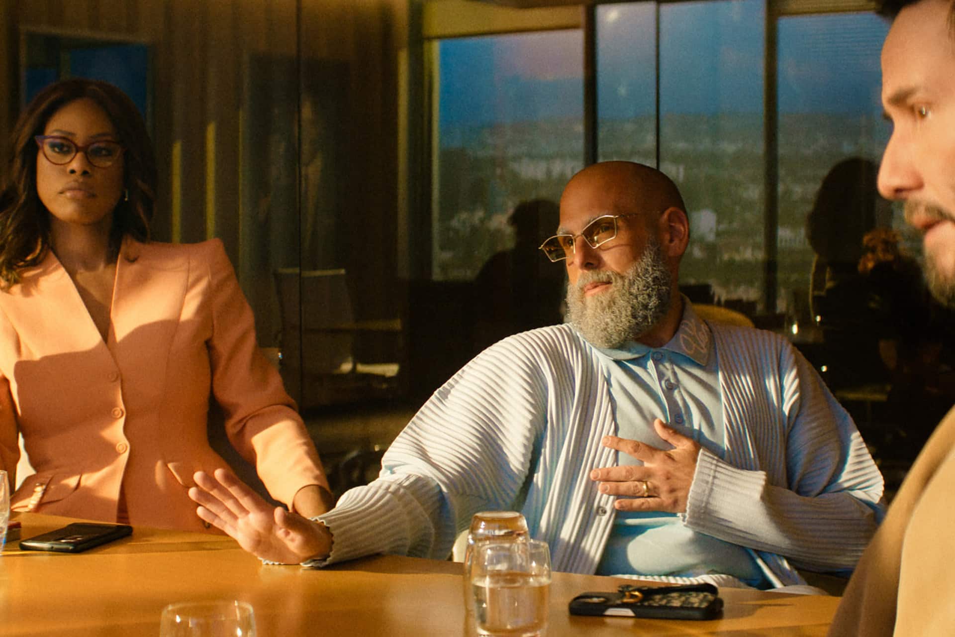 Three people sit around a conference table; one gestures with a raised hand and the other on their chest, sporting sunglasses and a beard, while another looks on seriously—capturing the suspense of an Outcome Trailer moment.