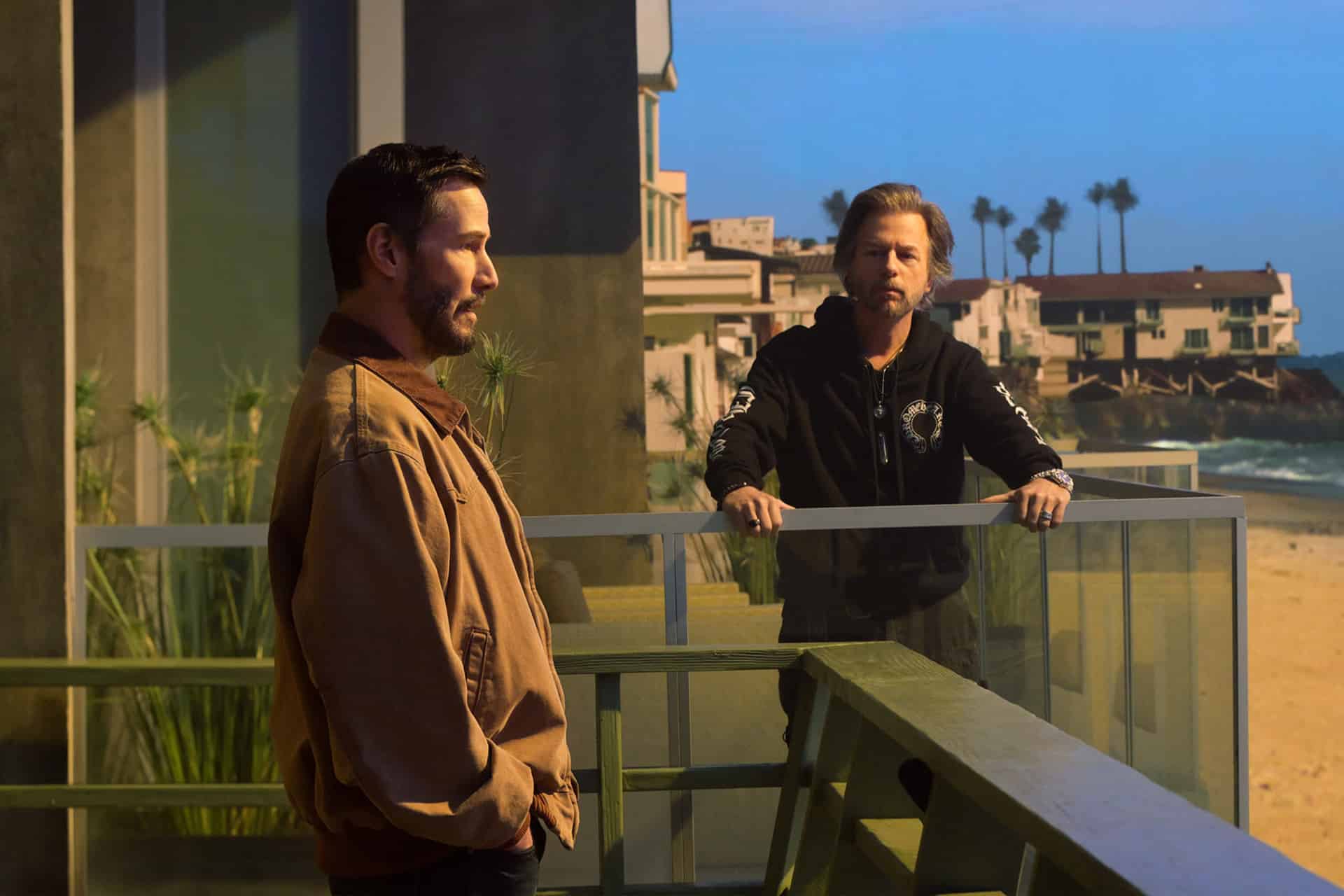 Two men stand on a beachfront balcony in this Outcome Trailer; one faces forward in a brown jacket, while the other in a black hoodie leans on the glass railing. Palm trees and beach houses stretch out in the background.