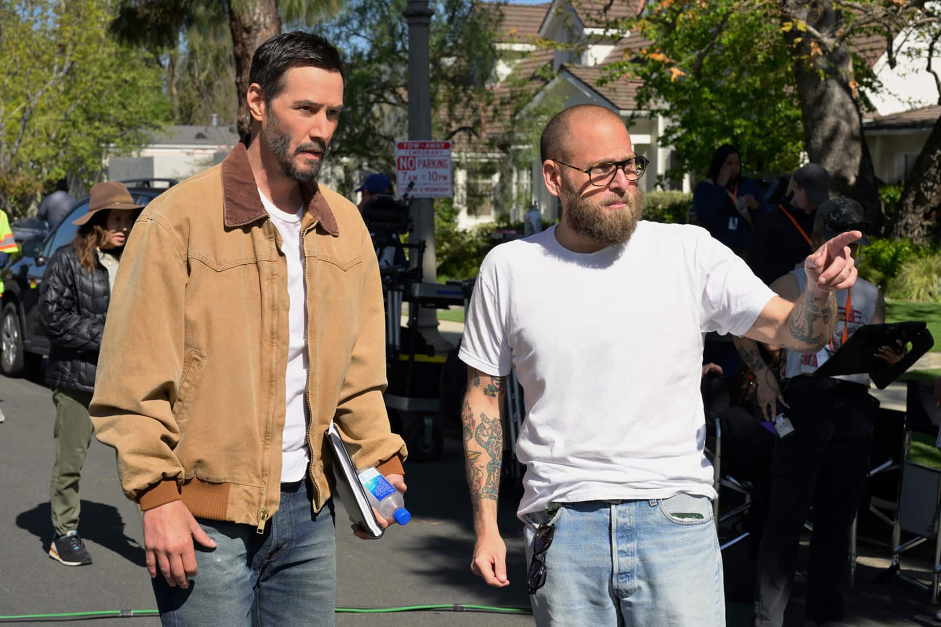 Two men stand outdoors on a film set, collaborating on the Outcome Trailer. One wears a tan jacket and holds a notebook, while the other, in a white t-shirt with tattoos, gestures forward. Crew members and equipment are visible in the background.