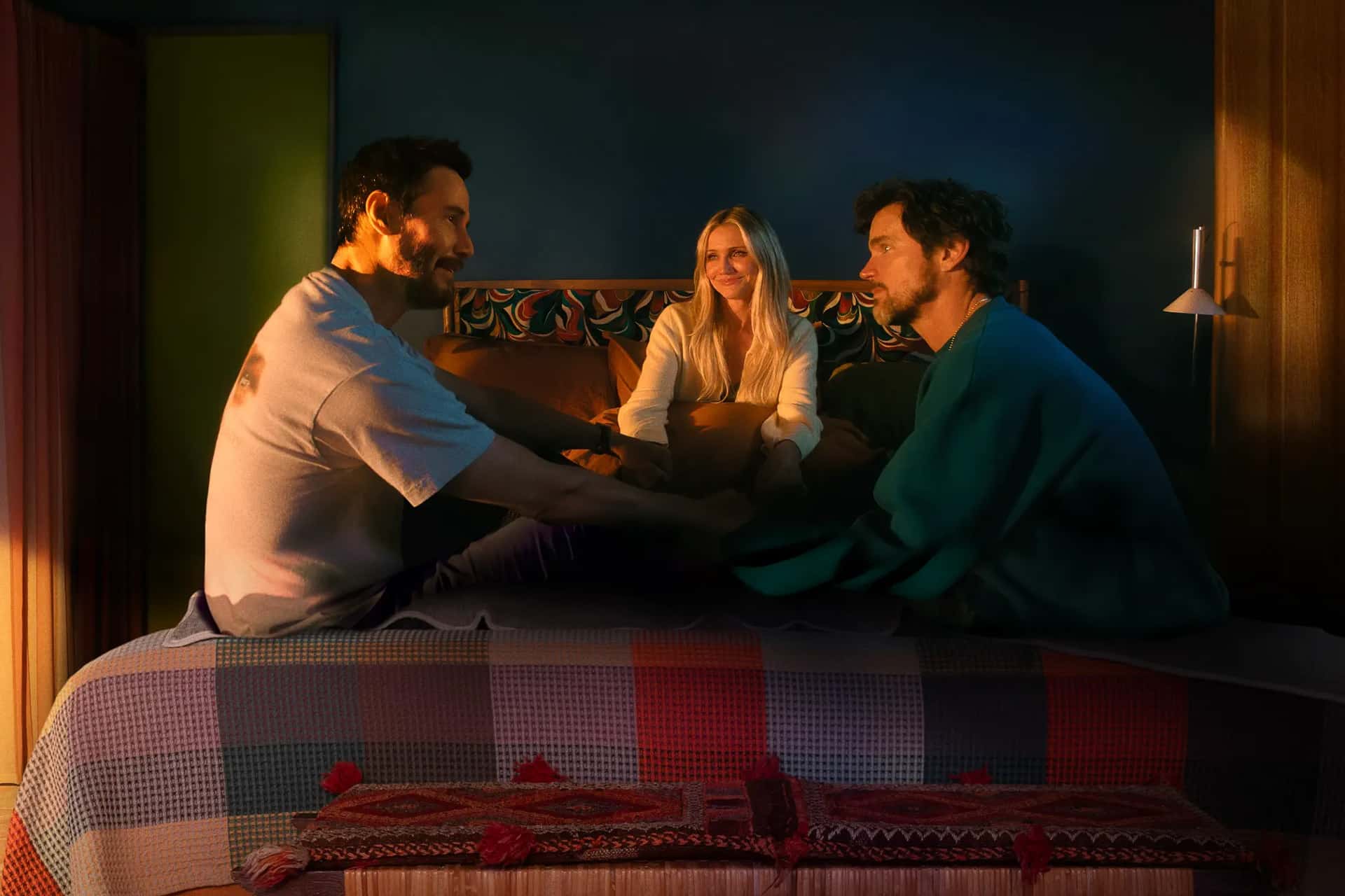 Three people sit together on a bed in a warmly lit room. Two men, holding hands, face each other while a woman smiles between them. The patchwork quilt and colorful decor create an inviting scene, echoing the heartfelt connections in the Outcome Trailer.