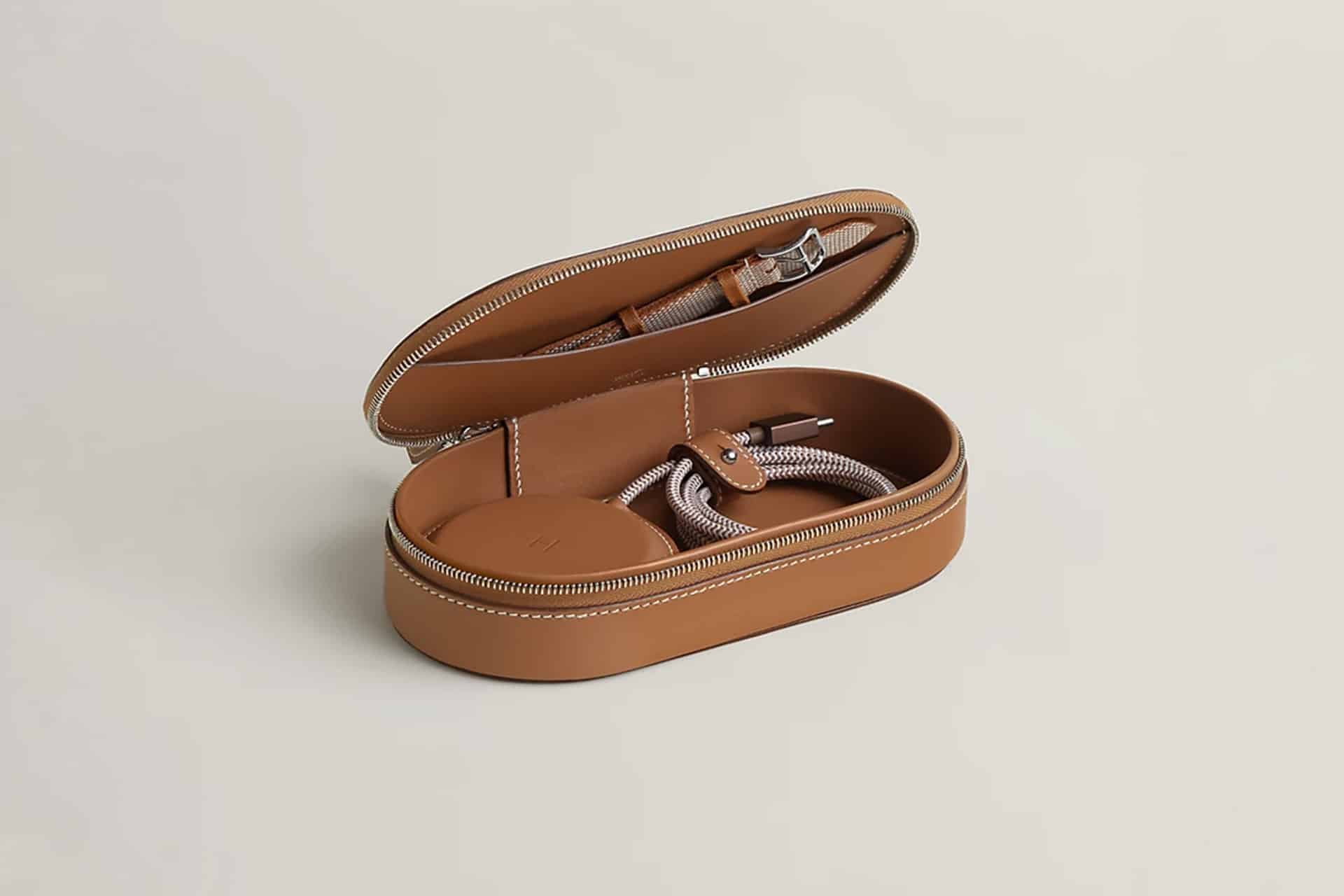 A tan leather zippered case is open, revealing a watch, a Hermès MagSafe Charger, and a braided charging cable neatly stored inside against a light background.