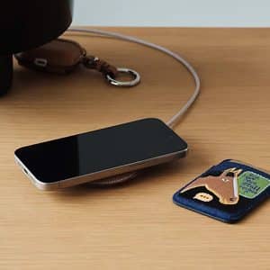 A smartphone rests on a wooden surface beside a blue cardholder featuring a horse illustration, a keychain with a brown leather strap, and an elegant Hermès MagSafe Charger.