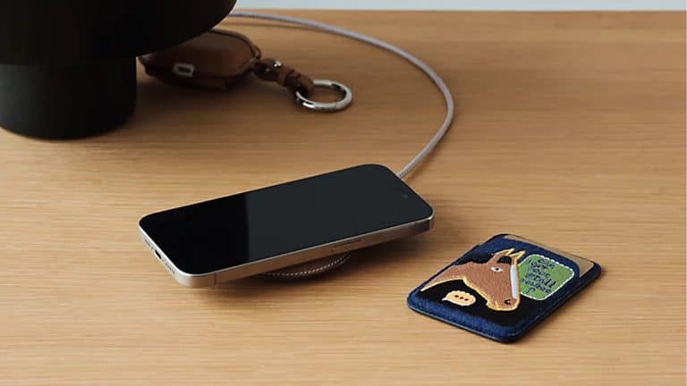 A smartphone rests on a wooden surface beside a blue cardholder featuring a horse illustration, a keychain with a brown leather strap, and an elegant Hermès MagSafe Charger.