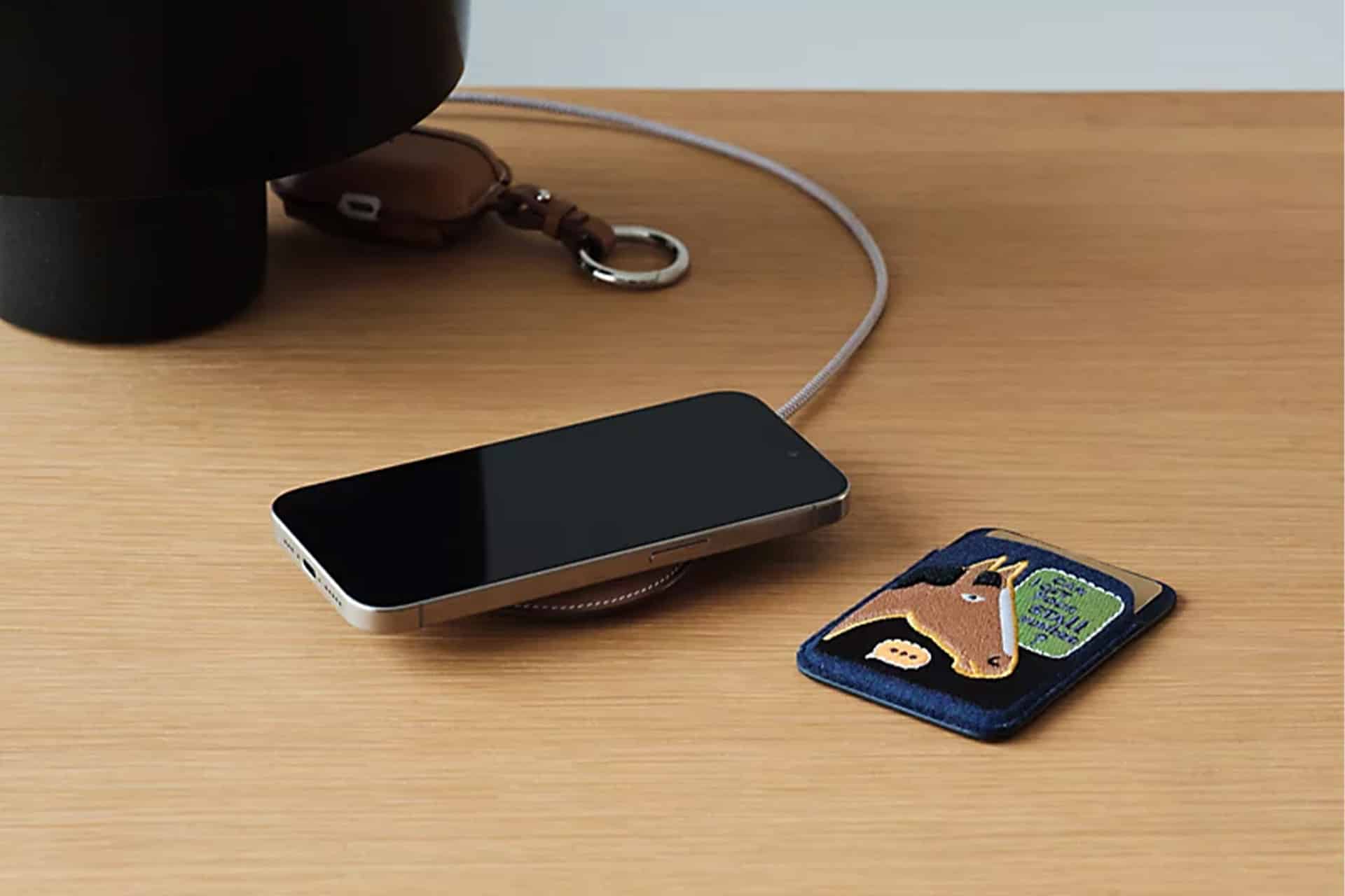 A smartphone rests on a wooden surface beside a blue cardholder featuring a horse illustration, a keychain with a brown leather strap, and an elegant Hermès MagSafe Charger.