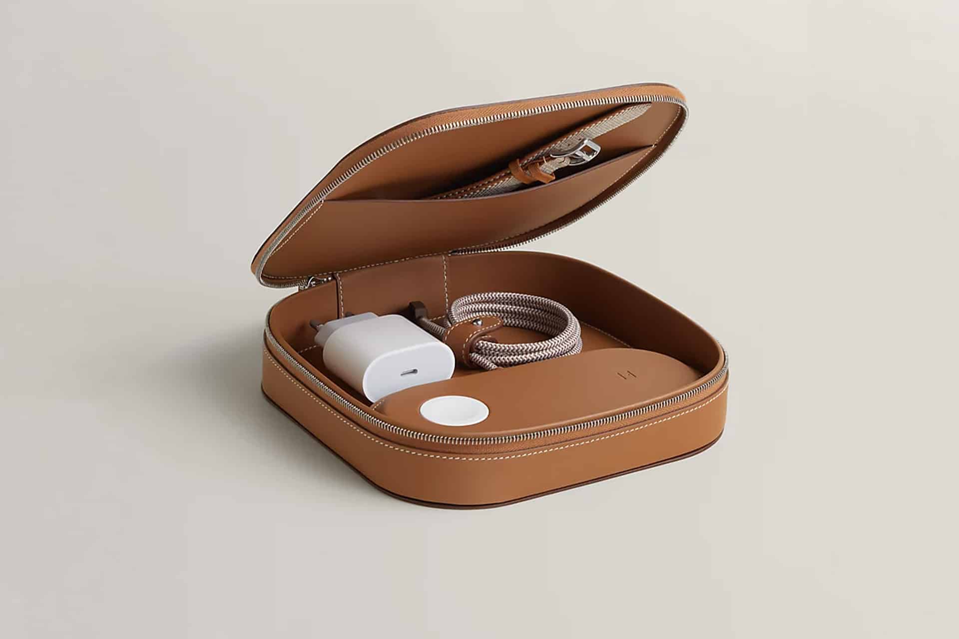 A tan leather zippered case is open, revealing a neatly coiled charging cable, a white power adapter, and a round Hermès MagSafe Charger inside on a neutral background.