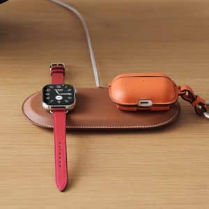A brown leather Hermès MagSafe Charger holds a smartwatch with a red band and an orange wireless earbud case with a keyring, both resting on a wooden surface.