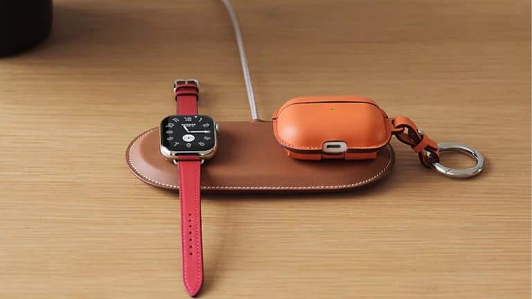 A brown leather Hermès MagSafe Charger holds a smartwatch with a red band and an orange wireless earbud case with a keyring, both resting on a wooden surface.