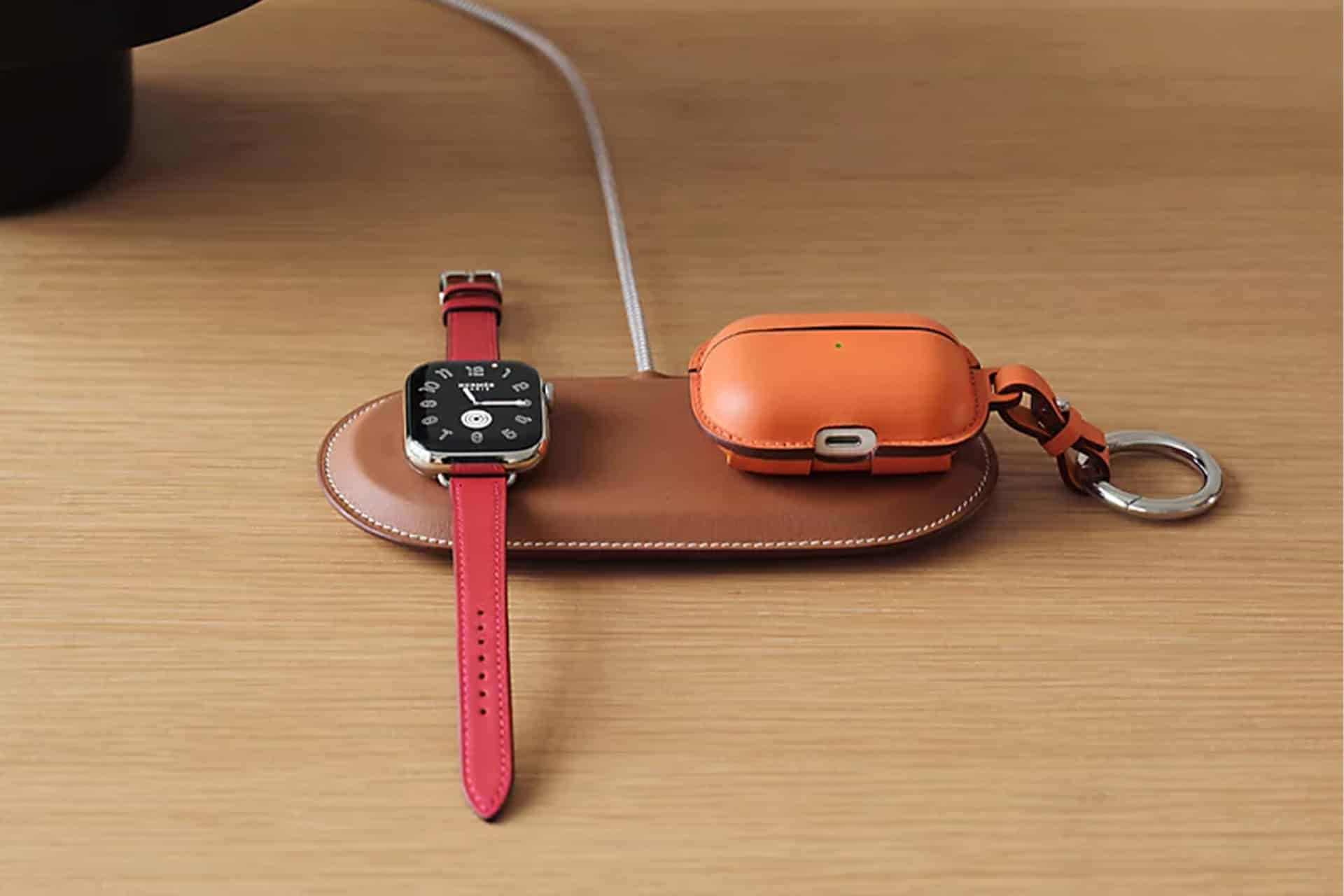 A brown leather Hermès MagSafe Charger holds a smartwatch with a red band and an orange wireless earbud case with a keyring, both resting on a wooden surface.