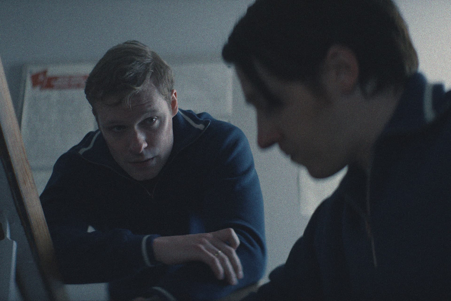 Two people in blue jackets are sitting indoors; one is looking intently at the other, who is slightly out of focus and facing downward. The dim lighting creates a serious, contemplative mood—reminiscent of scenes from the Star City Series on Apple TV.