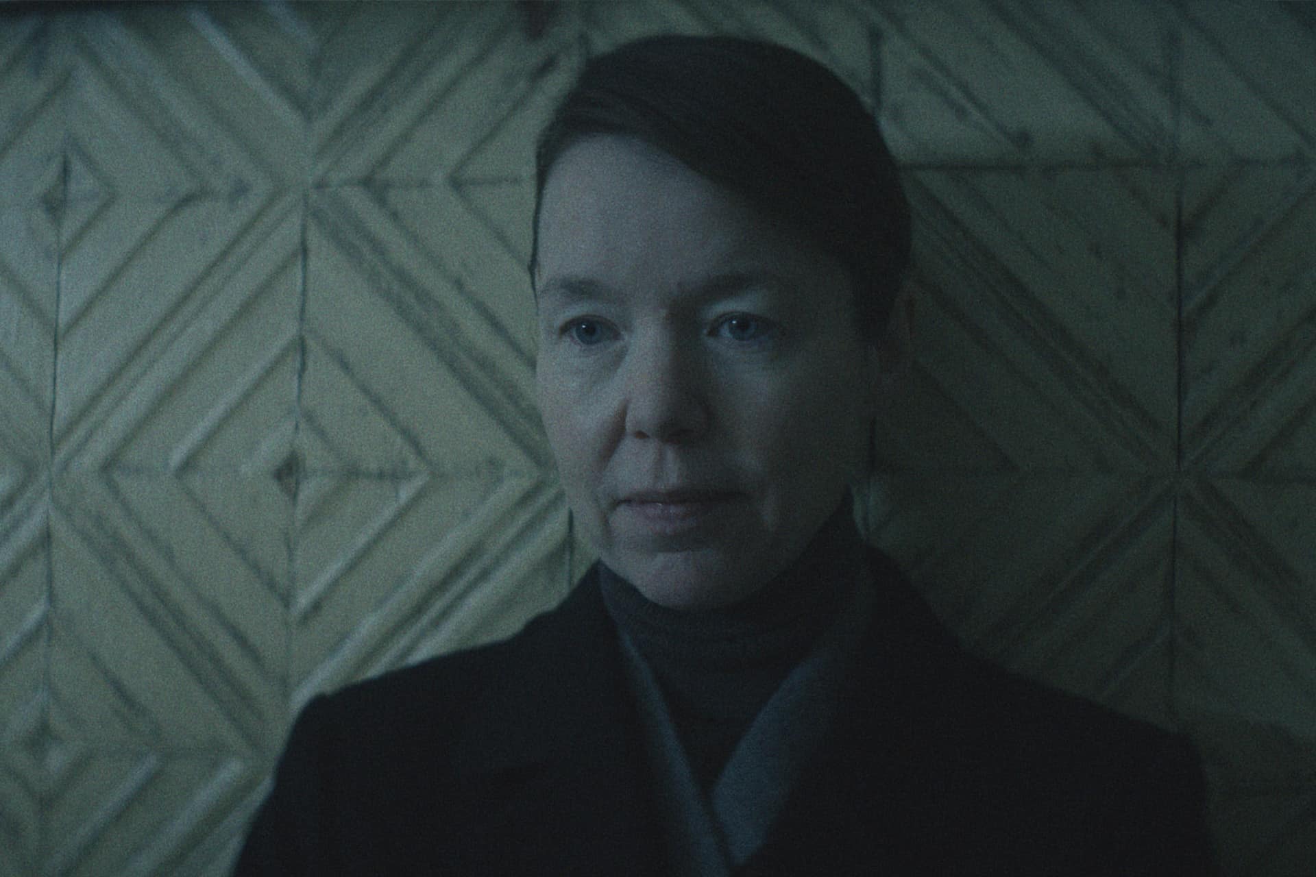 A person with short hair and a neutral expression stands against a wall with a geometric, patterned design. The moody lighting feels reminiscent of the ambiance in the Star City Series, casting soft shadows across their face and background.