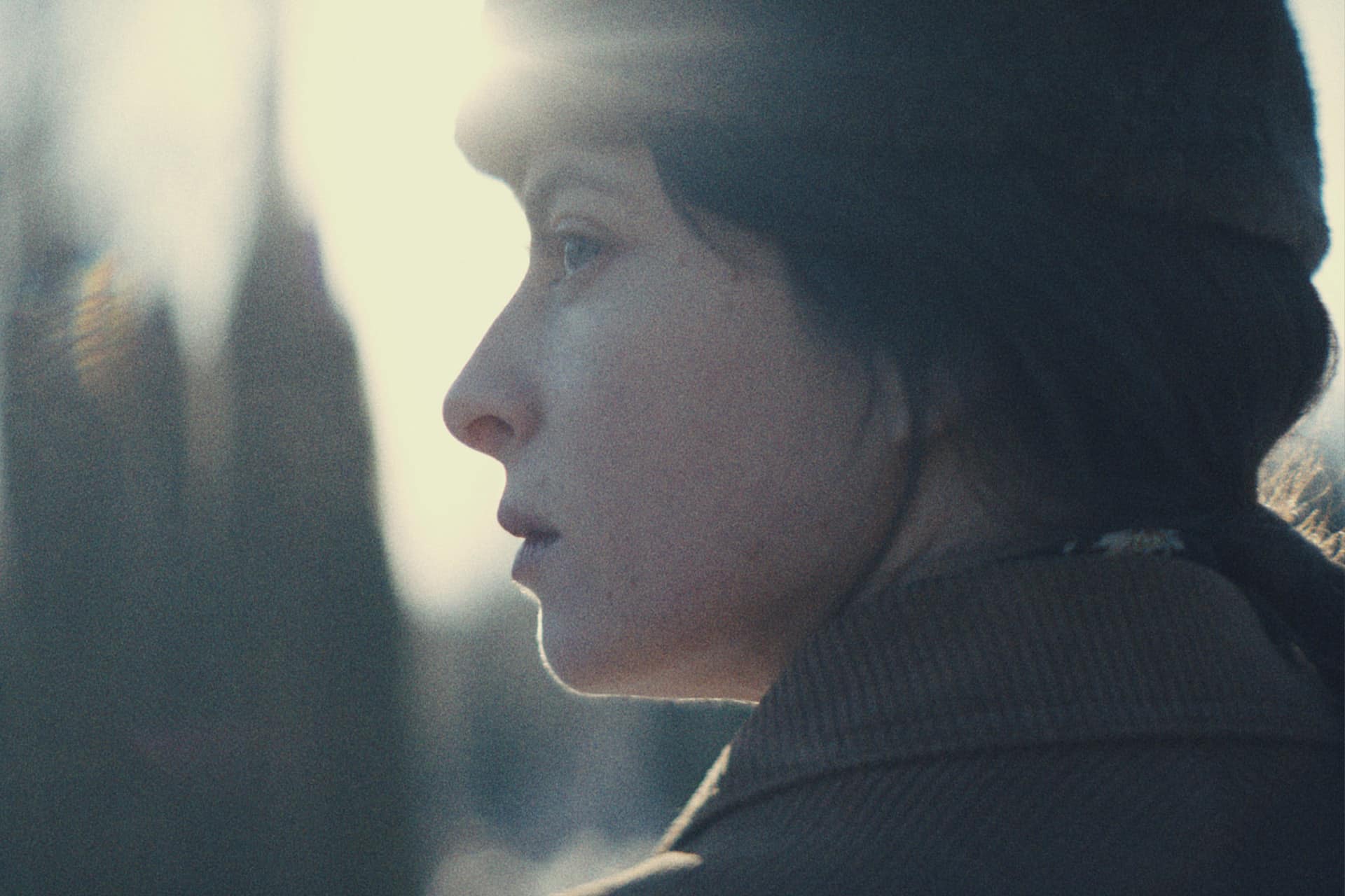 A person in profile wearing a dark hat and brown coat stands outdoors in soft, natural light, with sunlight creating a lens flare near their head and blurred trees in the background—evoking scenes from the Star City Series.
