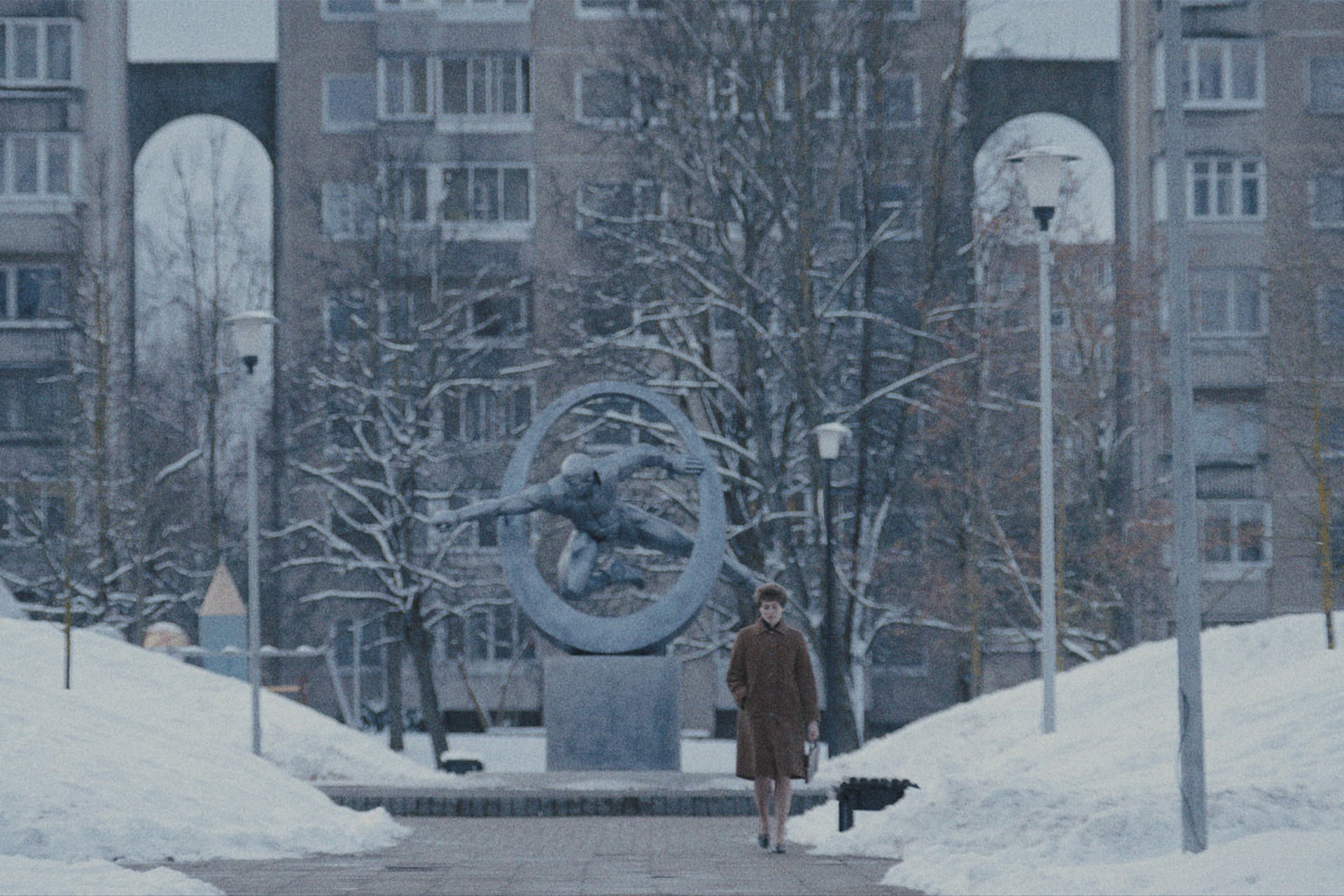 A person in a brown coat walks on a snowy path toward a large modern sculpture in Star City, with leafless trees and a tall apartment building—just like scenes from the Star City Series on Apple TV.