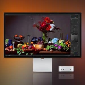 A sleek Apple monitor, tuned with Studio Display XDR settings, displays a vibrant still life of fruits, vegetables, and flowers beside a compact Apple desktop computer, all set against a gradient orange and brown background.