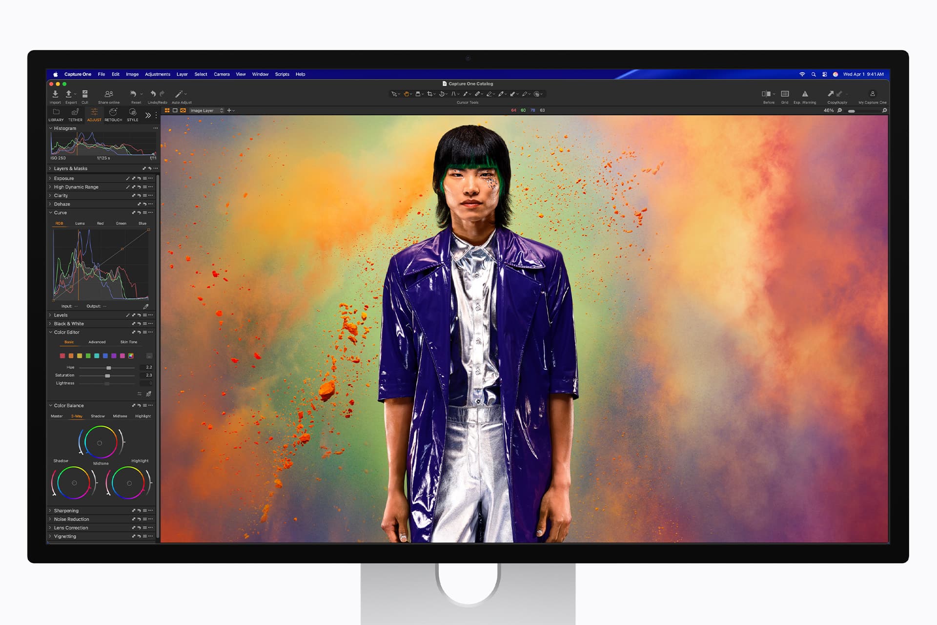 Studio Display XDR - A computer monitor displays an image-editing software with a photo of a person in a shiny purple jacket standing against a vibrant, colorful powder explosion background. Editing tools and color curves are visible on the screen.