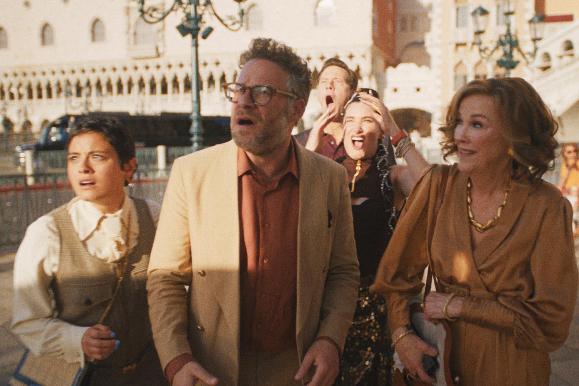 Five people stand closely together outdoors, all looking ahead with surprised or shocked expressions. Dressed in stylish clothing and illuminated by warm sunlight, they appear as if cast from an Apple TV The Studio series, with an ornate building in the background.