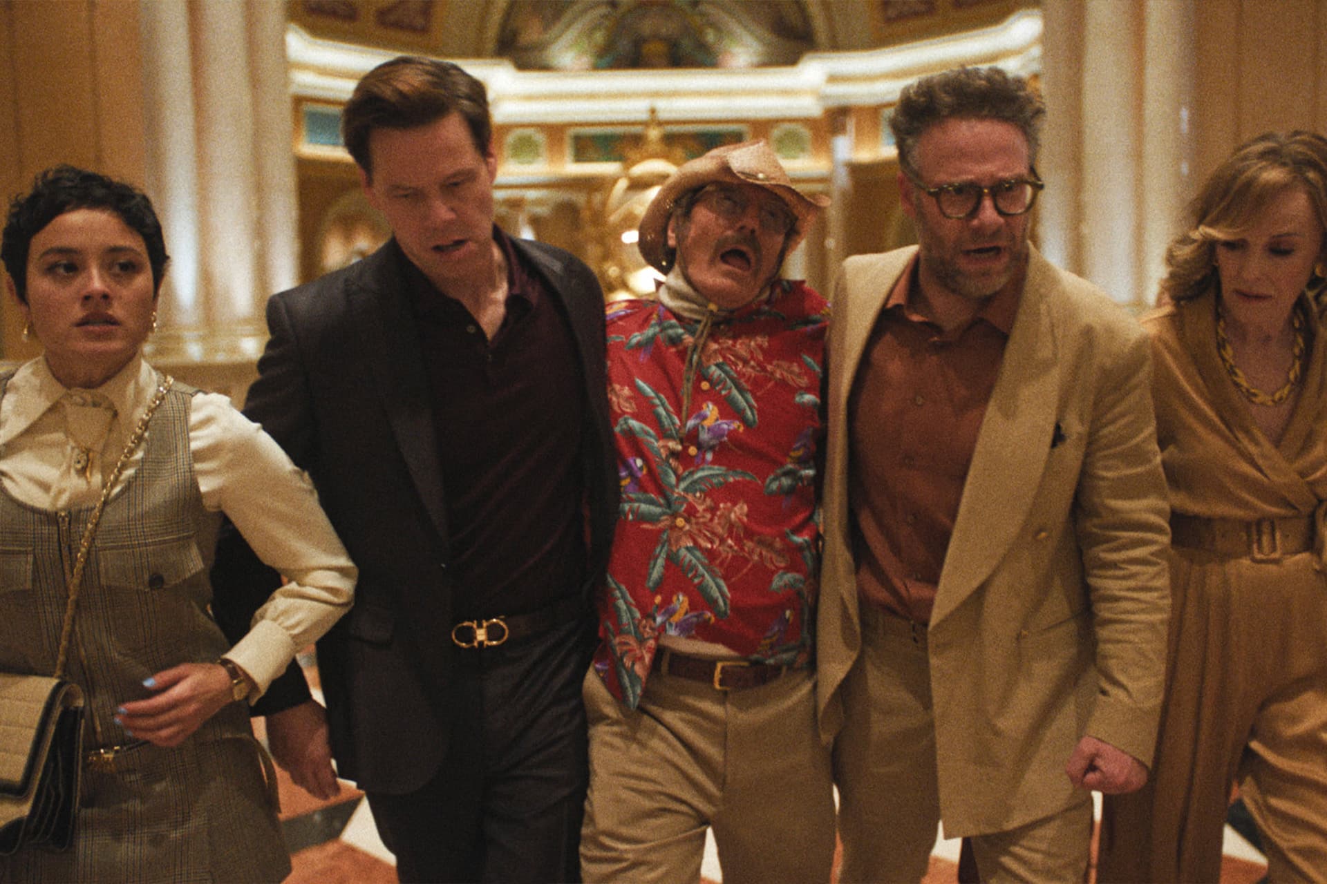 Five people walk together indoors, supporting a man in the center who appears unwell. The group looks concerned as they move through an ornate hallway featured in Apple TV The Studio, with columns and golden decorations.