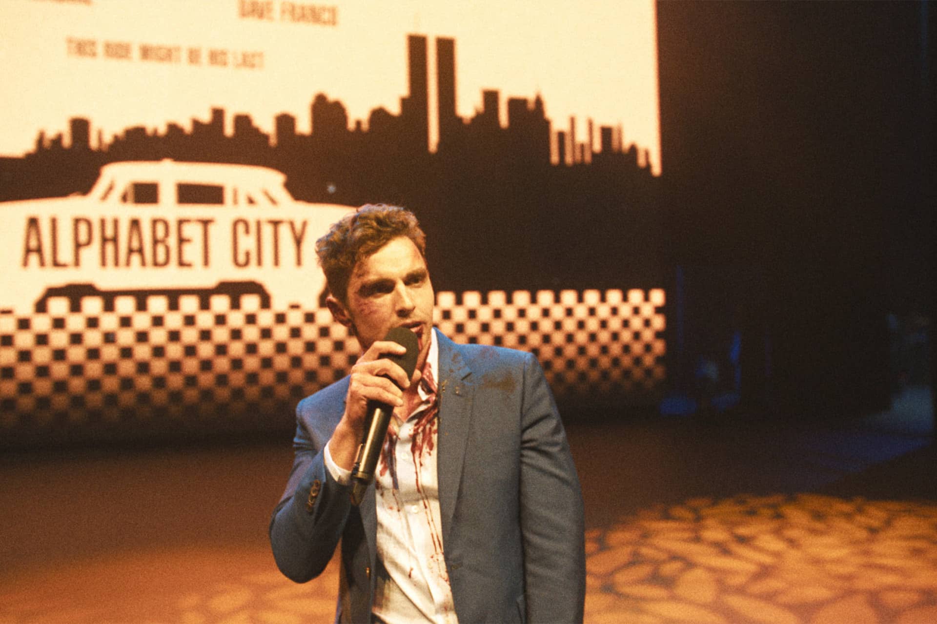 A man in a blue suit speaks into a microphone on stage, with a backdrop featuring a city skyline, the words "ALPHABET CITY," and an illustrated taxi. Warm lighting casts patterned shadows on the floor at Apple TV The Studio.