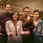 Four people stand together indoors, dressed in stylish, modern outfits, appearing engaged and expressive in an office or detective agency setting—possibly a scene from Apple TV The Studio, with beige walls and a desk in the background.