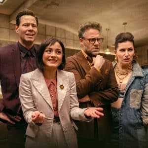 Four people stand together indoors, dressed in stylish, modern outfits, appearing engaged and expressive in an office or detective agency setting—possibly a scene from Apple TV The Studio, with beige walls and a desk in the background.