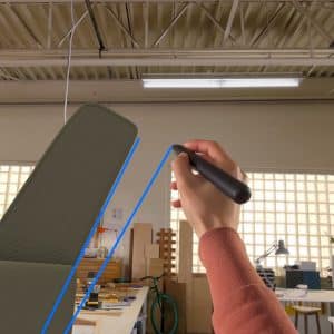 A person’s hand holding a VR controller uses Vision Pro Gestures to draw a blue line in a mixed reality workspace, with a desk, chair, bicycle, and office supplies visible in the background.