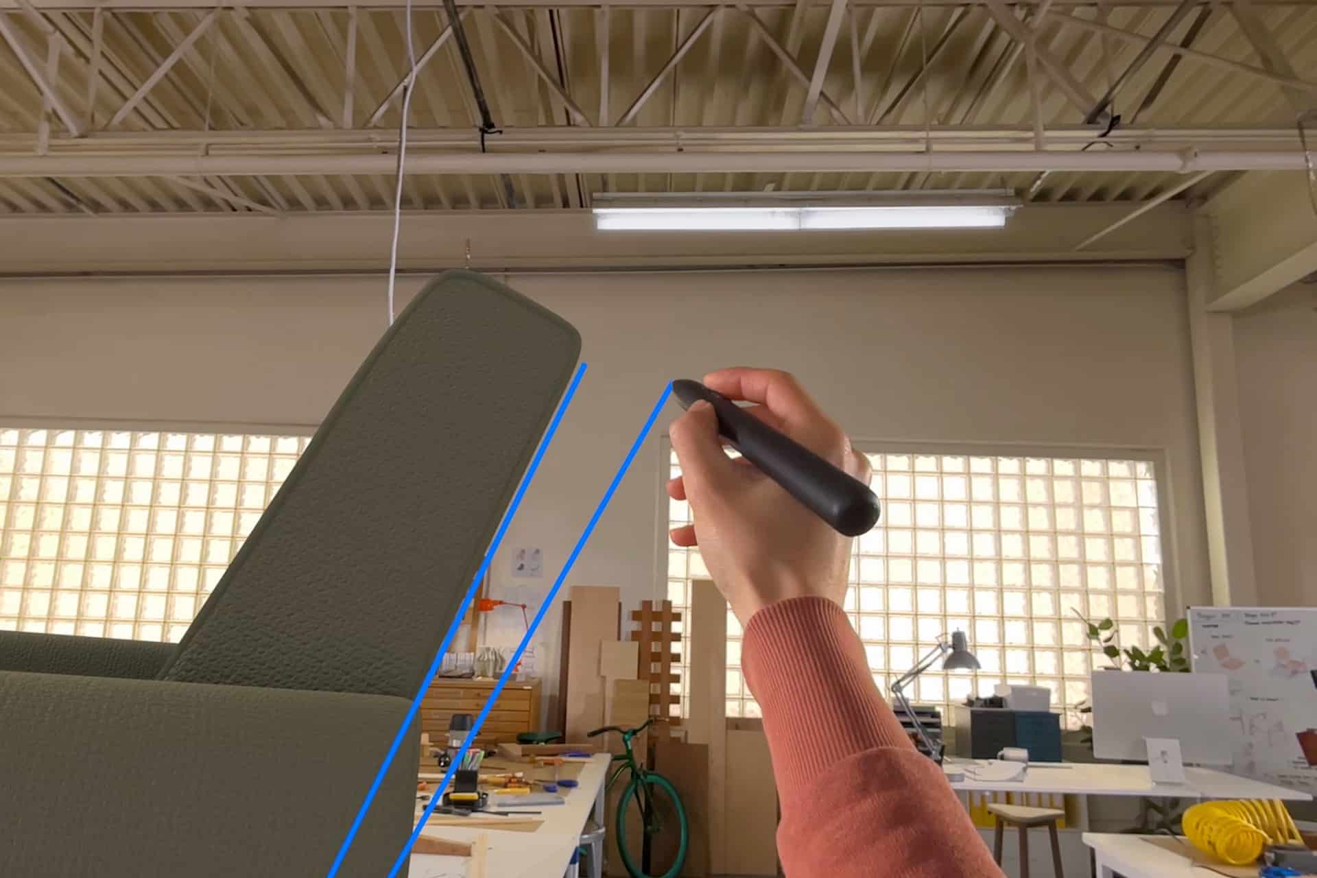 A person’s hand holding a VR controller uses Vision Pro Gestures to draw a blue line in a mixed reality workspace, with a desk, chair, bicycle, and office supplies visible in the background.