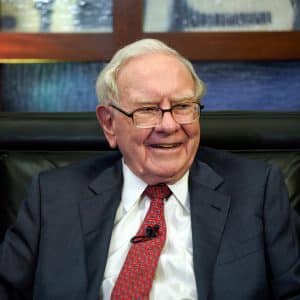 An elderly man with gray hair and glasses, wearing a dark suit, white shirt, and red patterned tie, sits smiling in a black leather chair. A microphone is clipped to his lapel as he discusses Warren Buffett’s investment in Apple stocks. - Warren Buffett Apple stocks