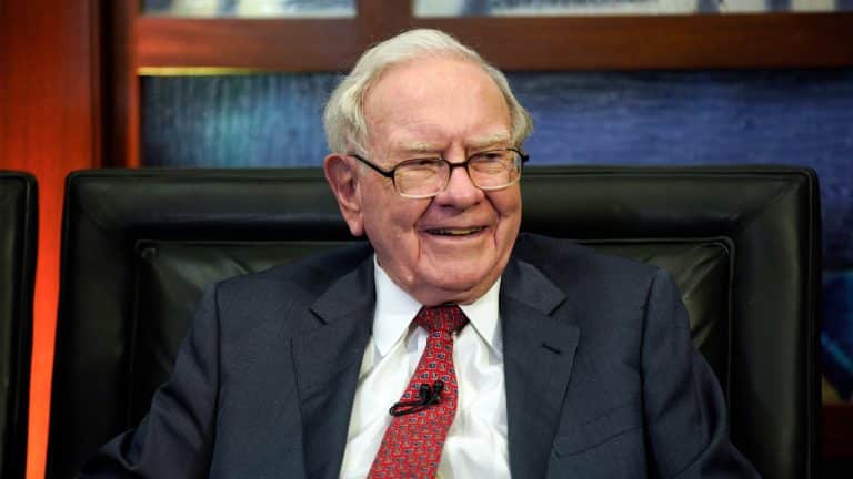 An elderly man with gray hair and glasses, wearing a dark suit, white shirt, and red patterned tie, sits smiling in a black leather chair. A microphone is clipped to his lapel as he discusses Warren Buffett’s investment in Apple stocks. - Warren Buffett Apple stocks
