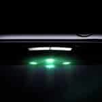 A close-up, low-light image of the underside of a smartwatch emitting green light from its sensors, highlighting its sleek metallic finish and side buttons, showcases the watch’s focus on oxygen accuracy.