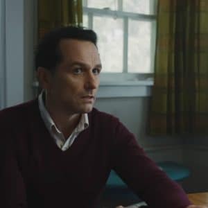 A man with short dark hair, wearing a maroon sweater over a collared shirt, sits at a wooden table in a softly lit room with green plaid curtains and a frosted window—the cozy setting evokes the charm of the Widow’s Bay Series.