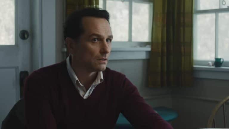 A man with short dark hair, wearing a maroon sweater over a collared shirt, sits at a wooden table in a softly lit room with green plaid curtains and a frosted window—the cozy setting evokes the charm of the Widow’s Bay Series.