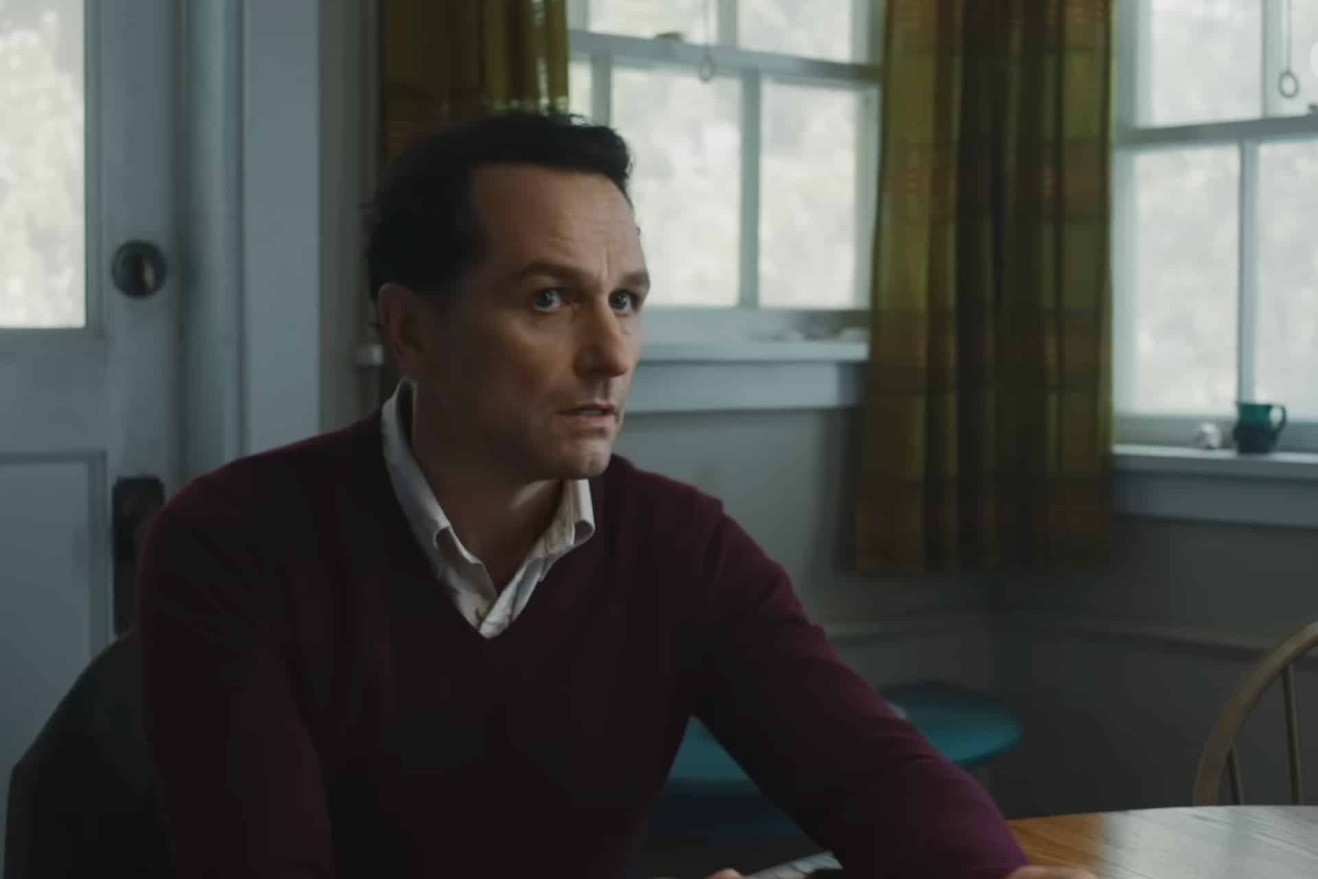 A man with short dark hair, wearing a maroon sweater over a collared shirt, sits at a wooden table in a softly lit room with green plaid curtains and a frosted window—the cozy setting evokes the charm of the Widow’s Bay Series.