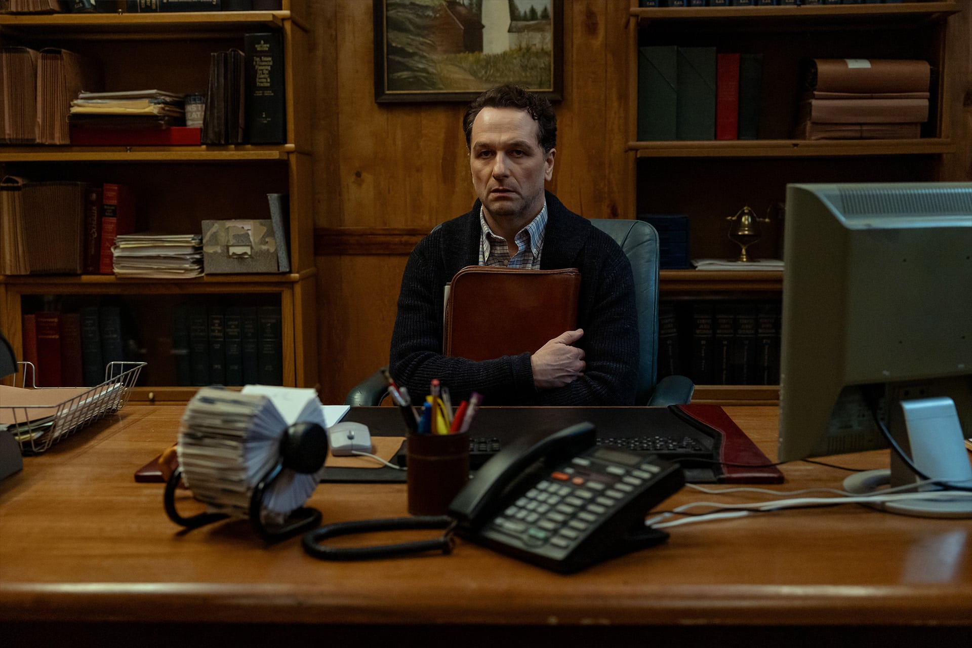 A man sits alone at a wooden desk in an office, holding a folder to his chest. The desk has office supplies, a phone, and a computer, while bookshelves filled with files and books—some from the Widow’s Bay Series—line the background.